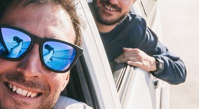 two young men leaning out from automobile windows, one with sunglasses that reflect his arm holding the camera taking the picture.